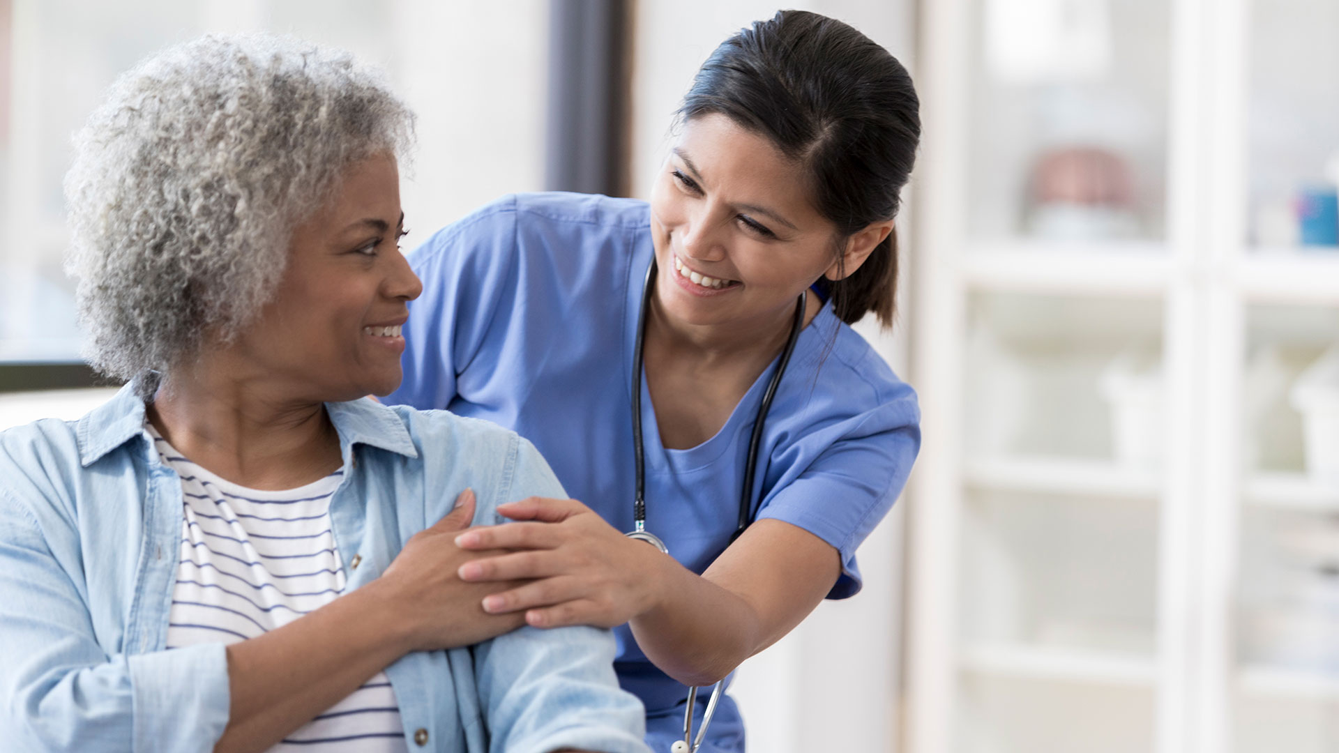 A patient sits in a chair while a doctor bends down to touch their hand.