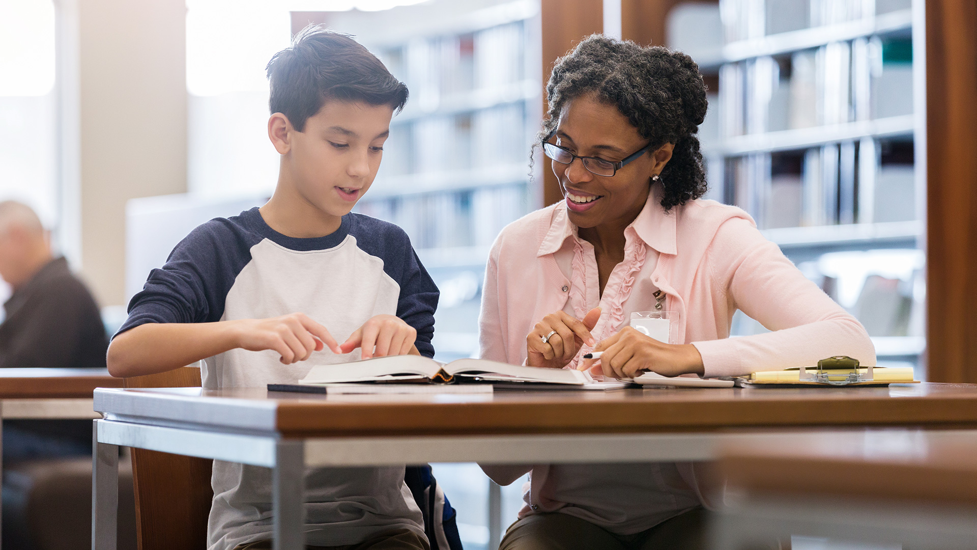 A teacher and a student sit at a library table and go over a book.