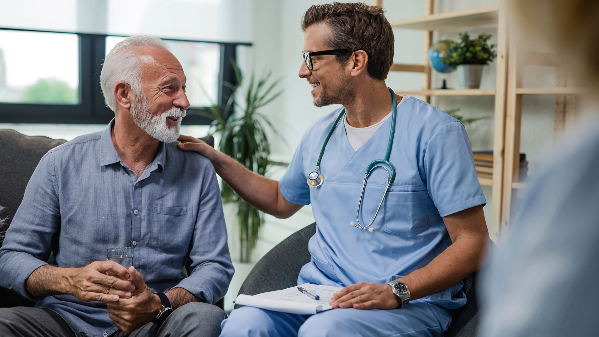 A doctor and a patient sit in chairs and talk while the doctor rests their hand on the patient’s shoulder.