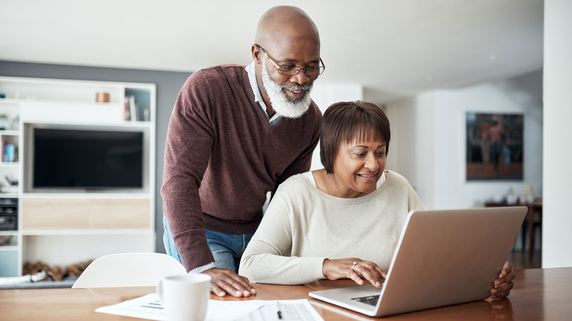 A person sits at a kitchen table while another person leans over them and look at a laptop.