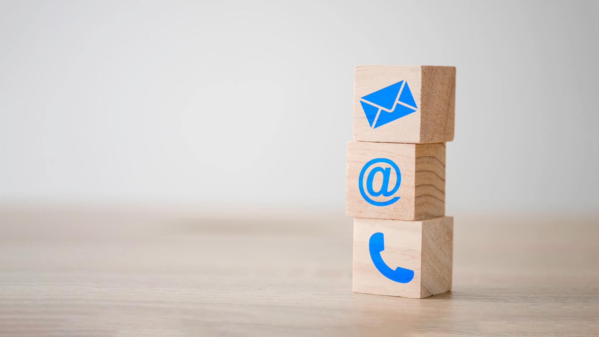 A stack of three wooden blocks, one with an envelope symbol, one with an at the rate symbol and one with a phone symbol, sit on a table.