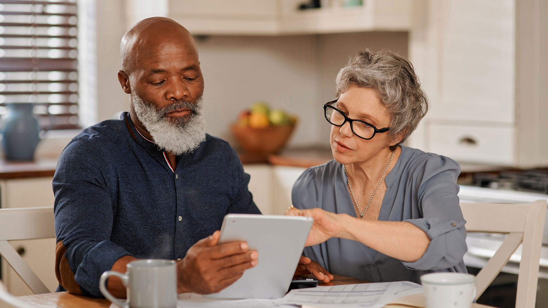 Two people sit at a kitchen table and look at a tablet.