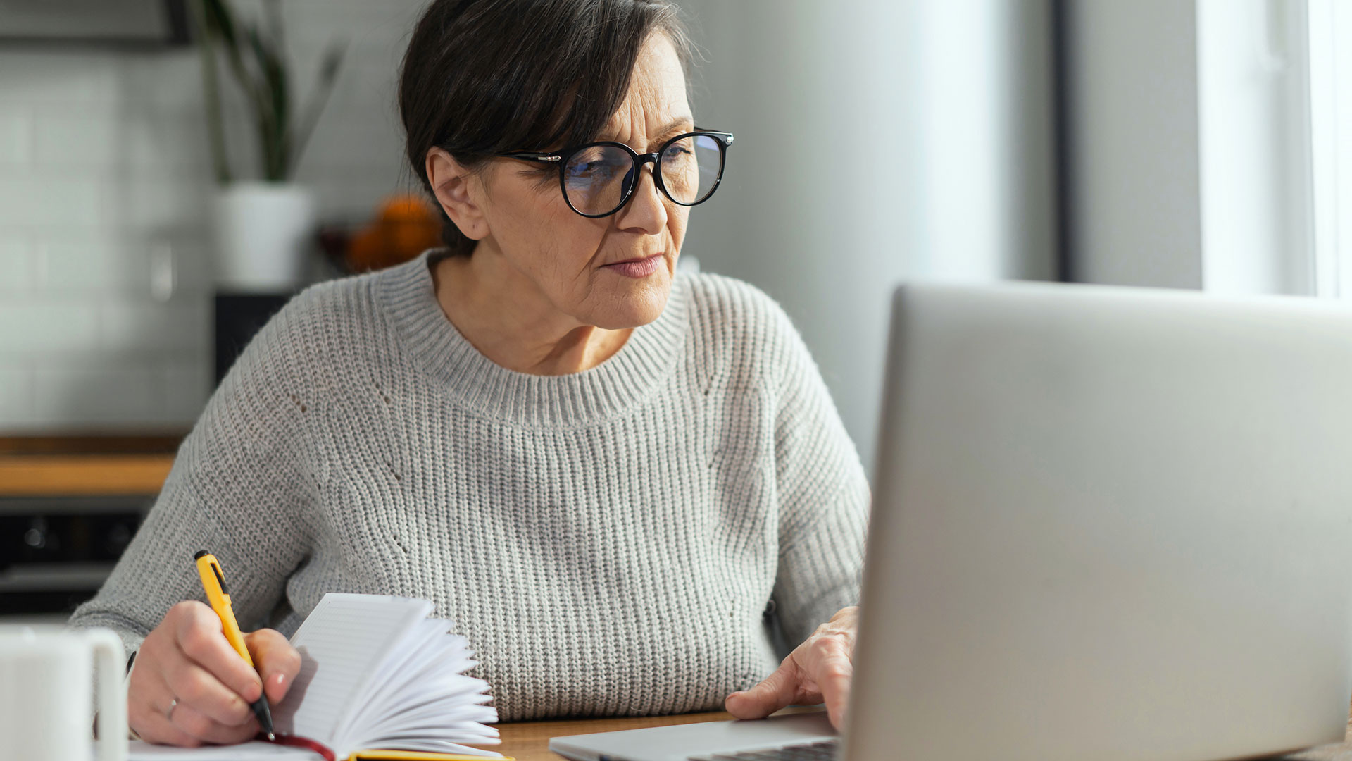 A person sits at a kitchen table and looks at a laptop.