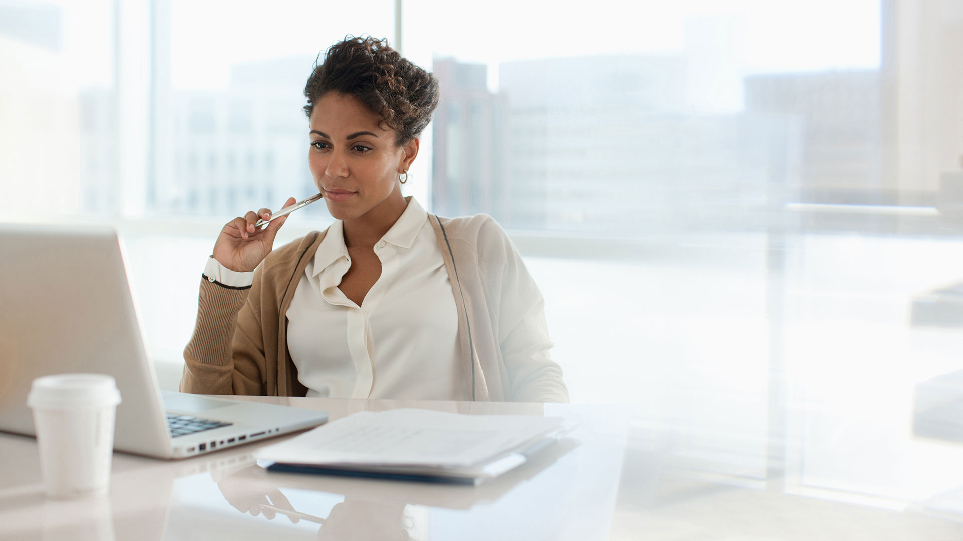 A person sits at a desk and looks at a laptop while they hold a pen to their cheek.