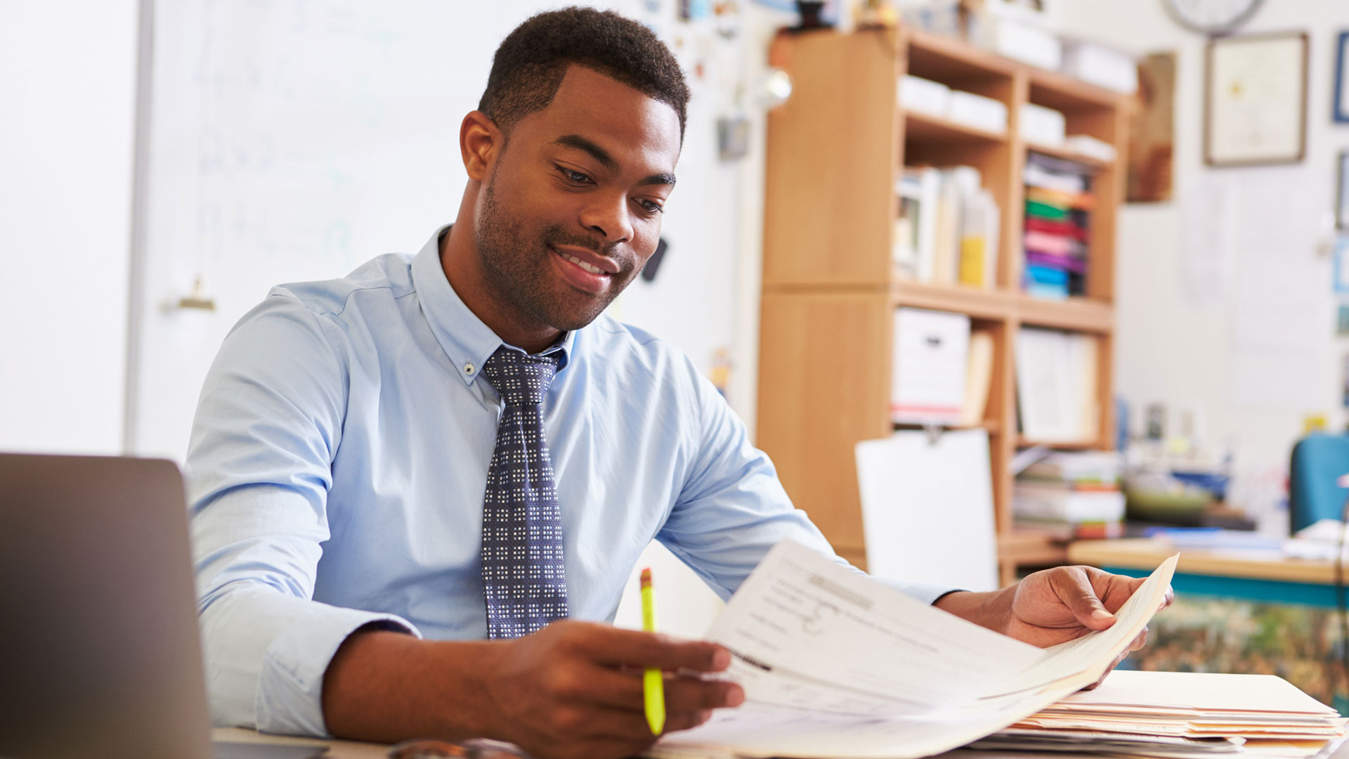 A teacher sits at a desk in a classroom and goes over papers.