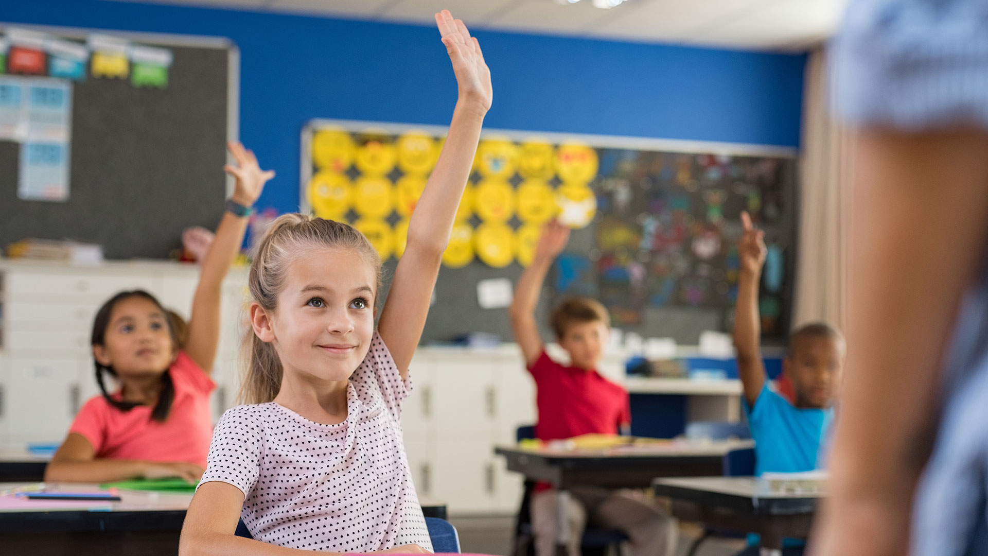 Children sit at desks in a classroom and raise their hands.